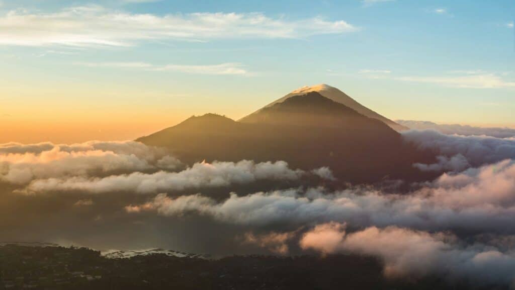 An image of Mount Batur sticking through the clouds in front of a yellow and blue sky 