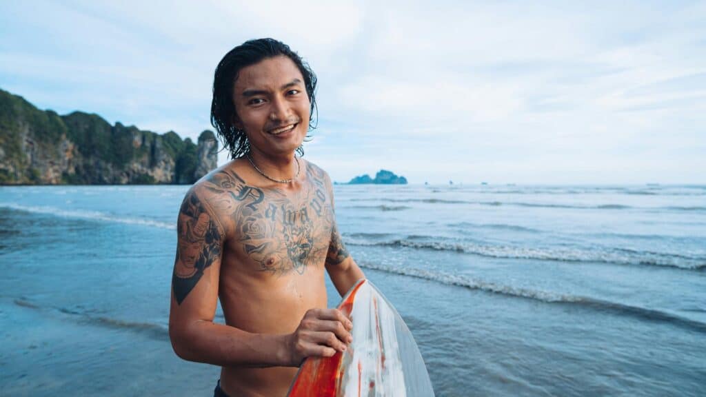 An image of a Indonesian man holding a surfboard on a beach in bali