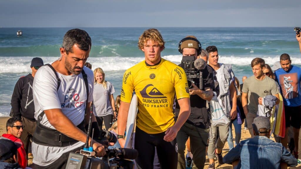 An image of john john florence leaving the ocean holding a surfboard he is surrounded by camera crew